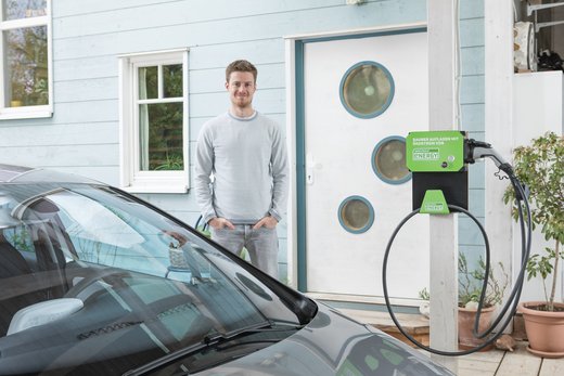 Ein Mann steht vor einem Haus neben einer Wallbox-Ladestation von Green Planet Energy. Im Vordergrund ist ein Auto zu sehen.