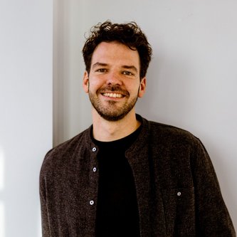 Ein Mann mit kurzen Haaren und Bart lächelt in die Kamera. Er trägt ein dunkles Hemd und darunter ein schwarzes Oberteil.