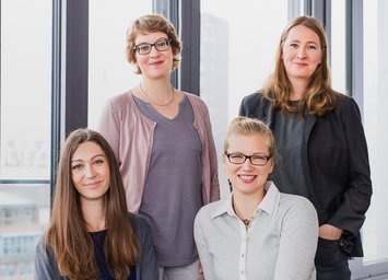 Vier Frauen vor einer modernen Fensterfront. Vorne Links sitzt eine Frau mit langen braunen Haaren, daneben eine blonde Frau mit Zopf und Brille. Hinter ihnen steht Links eine blonde, kurzhaarige Frau mit Brille. Rechts davon steht eine dunkelblonde langhaarige Frau. Alle lächeln in die Kamera.