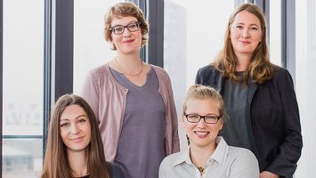 Vier Frauen vor einer modernen Fensterfront. Vorne Links sitzt eine Frau mit langen braunen Haaren, daneben eine blonde Frau mit Zopf und Brille. Hinter ihnen steht Links eine blonde, kurzhaarige Frau mit Brille. Rechts davon steht eine dunkelblonde langhaarige Frau. Alle lächeln in die Kamera.