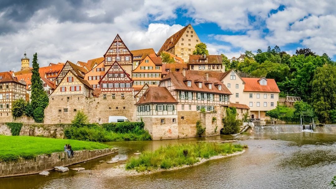 Fachwerkhäuser stehen an einer Flussböschung in einer historischen Stadt. Im Vordergrund sitzen zwei Menschen auf einer Wiese, während Wolken den Himmel bedecken.