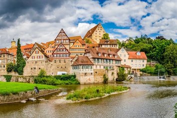 Fachwerkhäuser stehen an einer Flussböschung in einer historischen Stadt. Im Vordergrund sitzen zwei Menschen auf einer Wiese, während Wolken den Himmel bedecken.