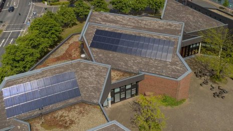 Flachdachgebäude mit Solarpanels, eingebettet in urbaner Umgebung mit Grünflächen und angrenzender Straße. Gewerbliches Gebiet im Hintergrund, begrenzt durch Verkehrsinfrastruktur. Nachhaltige Energieerzeugung in städtischem Kontext.