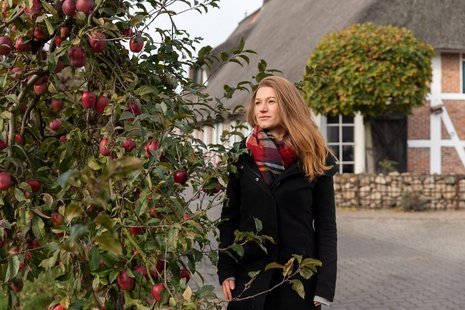 Eine Frau mit langen blonden Haaren steht vor einem Bauernhofhaus neben einem Apfelbaum mit roten Äpfeln.