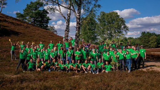 Große Gruppe Menschen mit grünen Green Planet Energy-T-Shirts steht in einer Heidelandschaft und lächelt bei Sonnenschein in die Kamera.