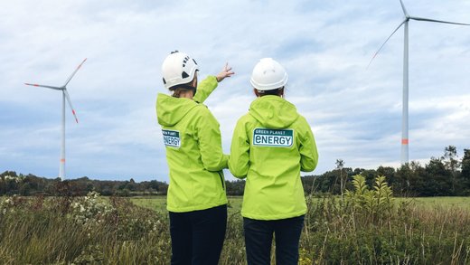 Zwei Personen mit Green Planet Energy Jacken stehen mit dem Rücken zur Kamera und blicken auf zwei Windkraftanlagen.