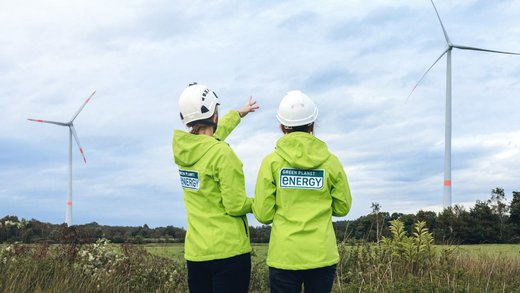 Zwei Personen mit Green Planet Energy Jacken stehen mit dem Rücken zur Kamera und blicken auf zwei Windkraftanlagen.