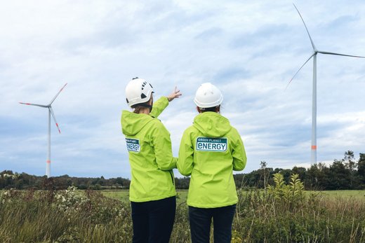 Zwei Personen mit Green Planet Energy Jacken stehen mit dem Rücken zur Kamera und blicken auf zwei Windkraftanlagen.