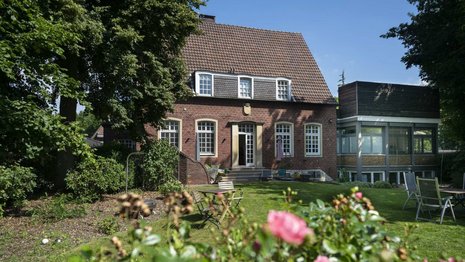 Ein zweistöckiges Backsteinhaus mit Giebeldach steht in einem Garten. Im Vordergrund blühen Rosen, umgeben von grüner Vegetation und Bäumen. Klarer, blauer Himmel im Hintergrund.