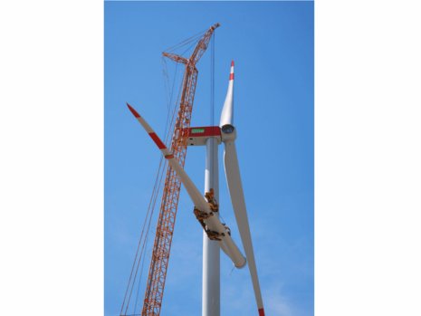 Ein Baukran befördert ein Rotorblatt nach oben zum unfertigen Windrad.