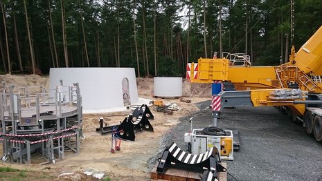 Baustelle einer Windkraftanlage mit einem Baufahrzeug auf der rechten Seite und einem Wald im Hintergrund.