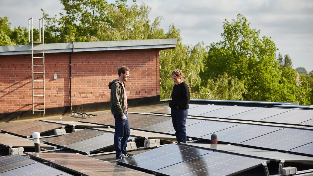 Zwei Personen in lässiger Kleidung stehen auf einem großen Flachdach, das komplett mit PV-Modulen bedeckt ist. Sie unterhalten sich lächelnd.