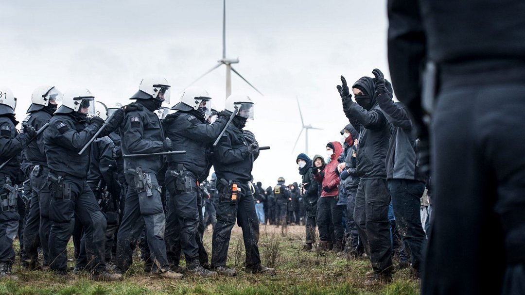 Eine Kette von Polizisten steht vor demonstrierenden Menschen auf einem Feld mit Windkraftanlage.
