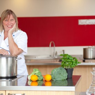 Mayoori Buchhalter, Geschäftsführerin von BioGourmetClub befindet sich in einer Küche und blickt in die Kamera. Auf der Theke liegen Gemüse und ein großer Topf.