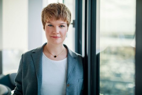 Frau mit kurzen blonden Haaren lehnt an einem Fenster und schaut in die Kamera
