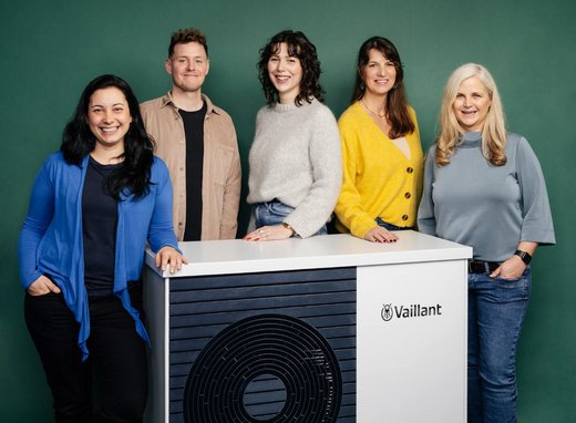 Eine Gruppe von 5 Menschen unterschiedlichen Aussehens stehen vor einem dunkelgrünen Hintergrund. Vor ihnen steht eine Wärmepumpe von Vaillant.