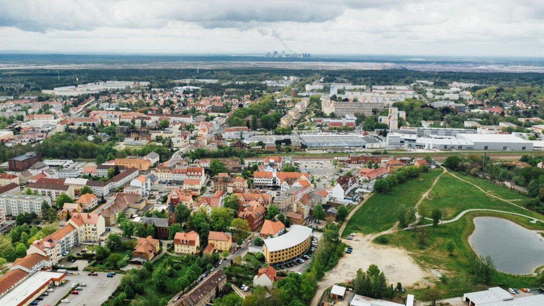 Luftaufnahme einer Stadt mit Wohnhäusern und Gewerbegebieten, flankiert von Grünflächen und einem kleinen See. Im Hintergrund zeigt sich ein bewaldeter Horizont unter wolkenverhangenem Himmel.