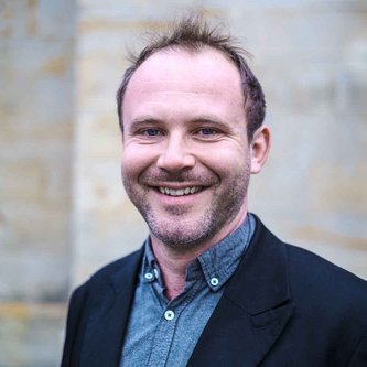 Ein Mann in blauem Hemd und schwarzen Sakko lächelt in die Kamera. Er hat einen Drei-Tage-Bart und dunkles Haar.