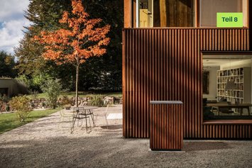 Modernes Haus mit Holzpaneelen als Außenwand. Links ist eine gelbe Sprechblase mit dem Text "Blogserie Teil 8" eingesetzt.