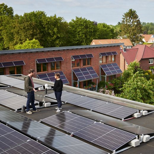 Eine Frau und ein Manns stehen auf dem Dach eines Mehrfamilienhauses, das mit PV-Modulen bedeckt ist. Sie unterhalten sich.