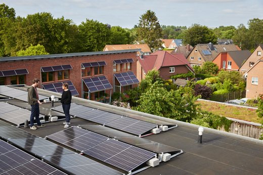 Eine Frau und ein Manns stehen auf dem Dach eines Mehrfamilienhauses, das mit PV-Modulen bedeckt ist. Sie unterhalten sich.