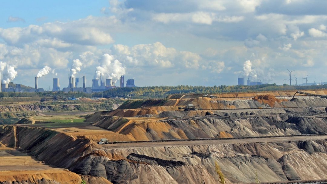 Braunkohletagebau im Vordergrund. Schwere Maschinen bewegen Erde. Im Hintergrund rauchende Industrieschornsteine vor bewaldetem Horizont. Wolken ziehen am Himmel entlang.