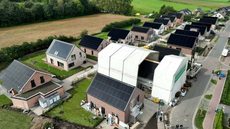Luftaufnahme eines Neubaugebiets mit Einfamilienhäusern, die Solarpaneele auf den Dächern haben. Arbeiter sind mit der Fertigstellung eines Hauses beschäftigt. Umgebung besteht aus Feldern und Wiesen.