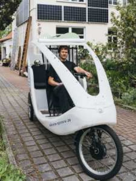 Ein Mann fährt ein dreirädriges Elektrofahrrad auf einem gepflasterten Weg vor einem Haus mit Solarpanelen. Das Elektrofahrzeug fördert nachhaltige Mobilität in städtischen Umgebungen.