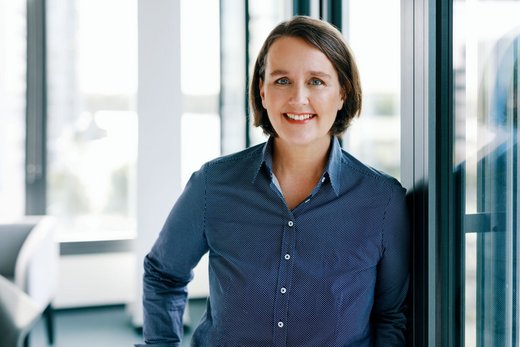 Frau mit kurzen braunen Haaren und gestreiftem Hemd lehnt in modernem Büro an einem Fenster.