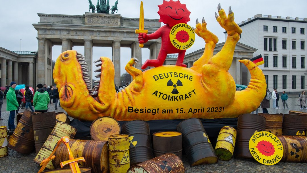 Ein gelber Dinosaurier aus Pappe liegt auf rostigen Fässern auf dem Rücken. Darauf steht "Deutsche Atomkraft - besiegt am 15.April 2023!"