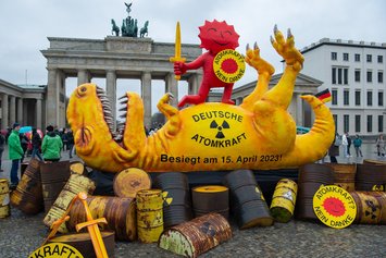Ein gelber Dinosaurier aus Pappe liegt auf rostigen Fässern auf dem Rücken. Darauf steht "Deutsche Atomkraft - besiegt am 15.April 2023!"