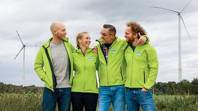Vier Personen mit einer Grüne Jacke von Green Planet Energy unterhalten sich. Im Hintergrund ein Feld und zwei Windräder.