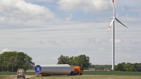 Fernaufnahme eines Feldes mit einer Windkraftanlage und einem Transportfahrzeug in der Mitte. Bäume stehen daneben.