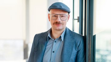 Porträt eines Mannes mit Brille und Schnurrbart, der eine Schiebermütze und ein blaues Sakko trägt. Er steht neben einem Fenster in einem hellen Innenraum