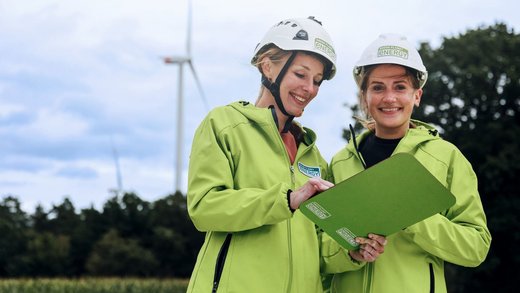 Zwei Frauen in saftgrünen Green Planet Energy Jacken vor einer Windkraftanlage. Sie halten ein Klemmbrett in der Hand und lächeln.