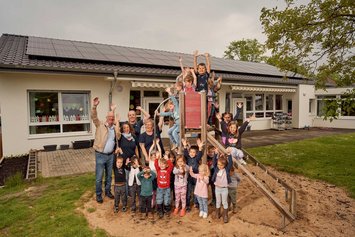 Eine Gruppe Kinder und einieg Erwachsene stehen vor und auf einem Klettergerüst, das im Sand vor einem flachen Haus steht