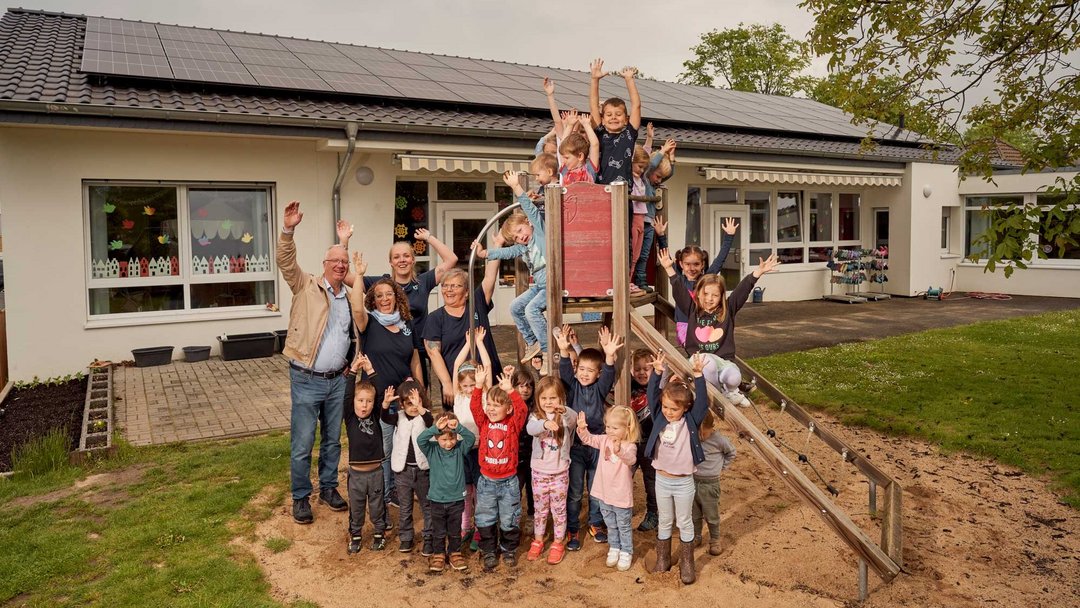 Eine Gruppe Kinder und einieg Erwachsene stehen vor und auf einem Klettergerüst, das im Sand vor einem flachen Haus steht