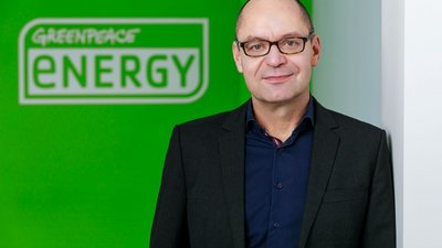 Ein Mann steht lächelnd in einem schwarzen Anzug vor einer Wand mit grünem Hintergrund. Auf der Wand steht: "GREENPEACE ENERGY".
