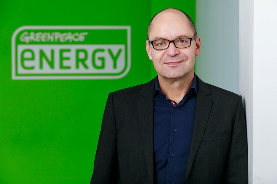 Ein Mann steht lächelnd in einem schwarzen Anzug vor einer Wand mit grünem Hintergrund. Auf der Wand steht: "GREENPEACE ENERGY".