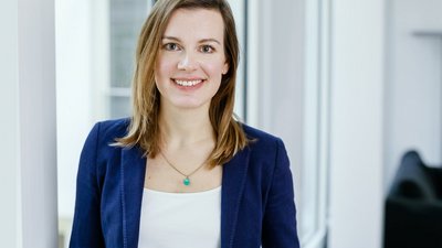 Eine Frau mit dunkelblonden Haaren steht vor einer großen Fensterfläche in einem Büro und lächelt in die Kamera.