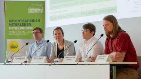 Drei Männer und eine Frau sitzen hinter einem Tisch mit Mikrofonen und Namensschildern vor sich; im Hintergrund wird etws an die Wand projiziert