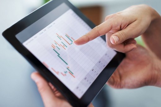 Hände halten ein Tablet, auf dem ein Balkendiagramm im Zeitverlauf in den Farben Grün und Organge abgebildet ist. Eine Hand zeigt darauf.