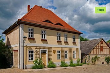 Älteres sandfarbenes Haus an einer Art Marktplatz. Das Haus hat kleine Fenster mit leeren Blumenkübeln davor. An der Hauswand sind Rankhilfen angebracht. Neben der Tür ist eine hölzerne beschriftete Tafel angebracht.