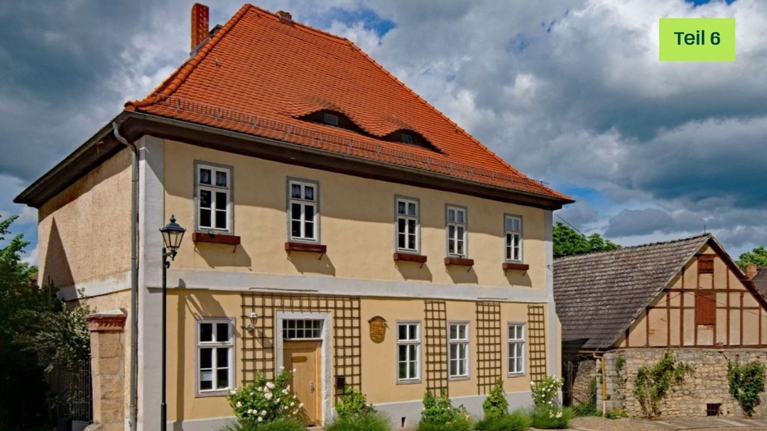 Älteres sandfarbenes Haus an einer Art Marktplatz. Das Haus hat kleine Fenster mit leeren Blumenkübeln davor. An der Hauswand sind Rankhilfen angebracht. Neben der Tür ist eine hölzerne beschriftete Tafel angebracht.