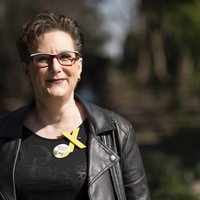 Eine Person steht lächelnd im Freien. Sie trägt eine schwarze Lederjacke und ein gelbes Schleifenband. Die Umgebung besteht aus Bäumen und unscharfem Hintergrund. An der Jacke ist ein Anstecker befestigt.