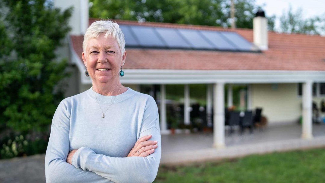 Eine Frau mit sehr kurzen weiß-grauen Haaren steht vor einem Haus mit PV-Anlage auf dem Dach. Sie trägt einen hellgrauen Pullover und lächelt mit verschränkten Armen in die Kamera.