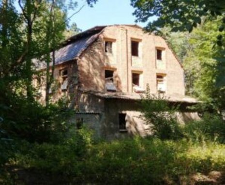 Ein verlassenes, zweigeschossiges Backsteingebäude mit kaputten Fenstern steht im Grünen, umgeben von dichtem Bewuchs und Bäumen, unter einem klaren blauen Himmel.