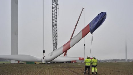 Die 3 Flügel der Windkraftanlage werden von einem Kran angehoben, während 2 Arbeiter den Vorgang überwachen.