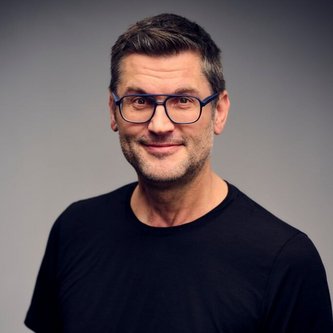 Eine Person mit Brille trägt ein schwarzes T-Shirt und lächelt in die Kamera. Der Hintergrund ist einfarbig grau, was das Porträt betont.