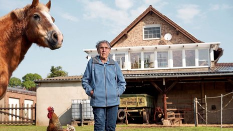 Eine Frau mit grauen kurzen Haaren steht vor einem Haus mit Solaranlage. Neben ihr stehen auf einer Wiese ein Pferd und ein Hahn.